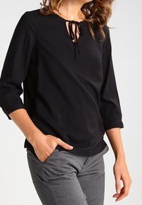 Blusa nera con scollatura rotonda e chiusura con lacci. Caratterizzata da maniche tre quarti e una vestibilità ampia, realizzata in tessuto morbido.