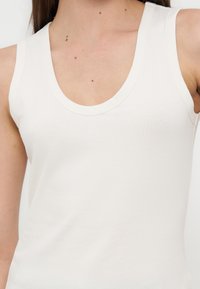 Γυναίκα που φοράει λευκό ριπ αμάνικο τοπ με στρογγυλή λαιμόκοψη, κοντινή λήψη στο πάνω μέρος του σώματος και τους ώμους.