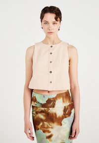 Korte beige linnen top met ronde halslijn en knopen aan de voorkant. Model draagt een kleurrijke, abstract-gepatroneerde rok met een middelhoge tailleband.