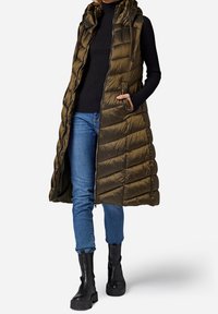 Długa oliwkowozielona kamizelka puchowa z kapturem, z pikowaną teksturą, zamkiem błyskawicznym i bocznymi kieszeniami, noszona na czarnym prążkowanym swetrze i niebieskich dżinsach.