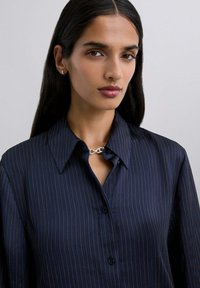 Camisa azul marino de botones con rayas verticales en naranja, con un collarina puntiagudo y un acento de cadena plateada en el escote.