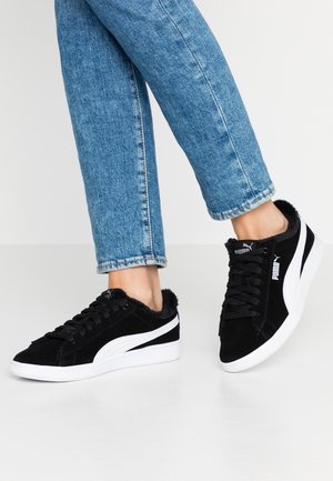 Fødder iført sorte Puma-sneakers med hvide såler og striber, kombineret med blå denimjeans mod en ensartet hvid baggrund.