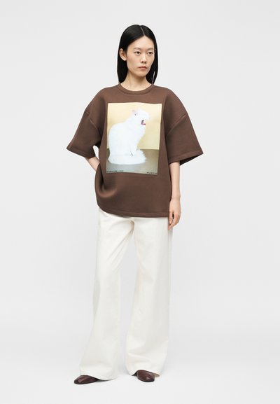 Μια γυναίκα στέκεται φορώντας ένα φαρδύ καφέ φούτερ με λευκό γραφικό γάτας, συνδυασμένο με φαρδιά λευκά παντελόνια και σκούρα καφέ παπούτσια.