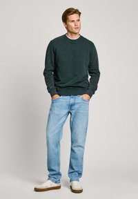Sudadera de cuello redondo verde oscuro con logo en relieve, combinada con jeans azules claros y zapatillas blancas con suelas marrones, sobre un fondo neutro.