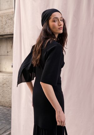 Woman with long brown hair wearing a black headscarf and black dress, standing sideways against a light pink fabric backdrop.