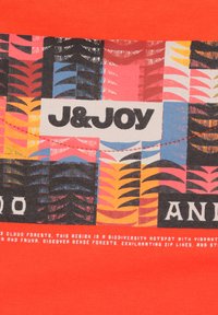 Rode katoenen t-shirt met een kleurrijk grafisch patroon van plantmotieven en de tekst "J&JOY." De tekst bevat feiten over biodiversiteit.