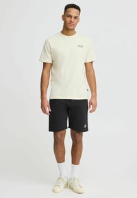T-shirt en coton crème à manches courtes et short noir. La T-shirt présente un petit logo sur le devant. Le modèle porte des chaussettes blanches et des baskets claires.