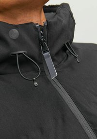 Sort jakke med høj krave, frontlynlås og snøre detaljer. Glat tekstur med mat finish og diskrete hardware accenter.