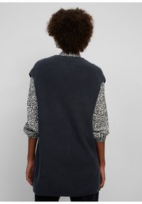 Veste tricotée gris foncé avec une surface texturée, dotée d'un col rond et d'emmanchures larges, portée par-dessus un chemisier léger à motif.