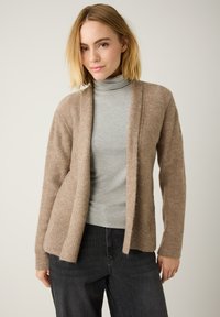 Hellbrauner Cardigan aus weichem Stoff, mit gerippten B�ündchen und offenem Frontdesign. Über einem grauen Rollkragenpullover und schwarzen Jeans getragen.