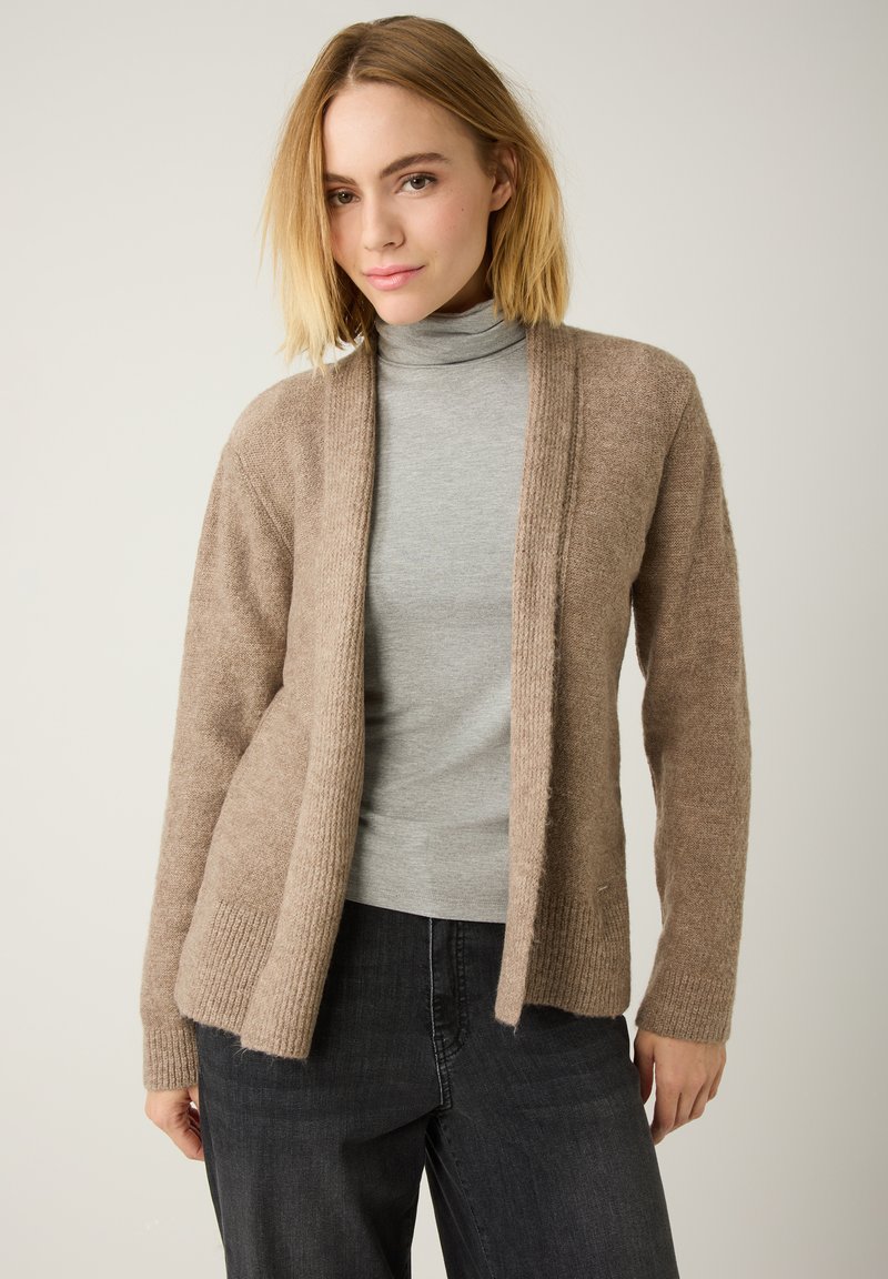 Hellbrauner Cardigan aus weichem Stoff, mit gerippten Bündchen und offenem Frontdesign. Über einem grauen Rollkragenpullover und schwarzen Jeans getragen.