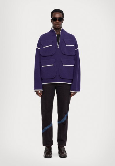 Pullover viola lavorato a maglia con accenti bianchi e numerose ampie tasche, abbinato a jeans scuri con un motivo diagonale blu e scarpe arrotondate.