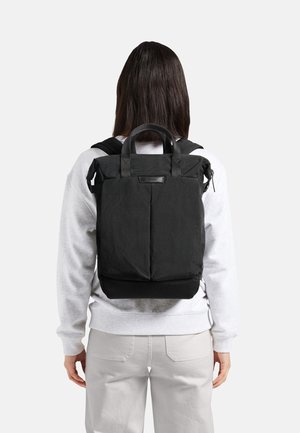 Sac à dos en tissu noir avec double poignée, bretelles rembourrées et forme rectangulaire. Présente une surface texturée et un design minimaliste.