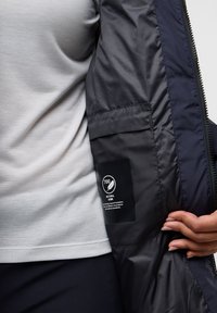 Binnenzijde van een marineblauwe pufferjack met een gladde textuur, voorzien van een label voor 700 fill power donsisolatie. Grijze T-shirt zichtbaar eronder.