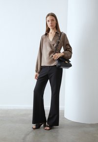 Brown satin wrap blouse with long sleeves, paired with black flared pants. Individual holds a textured black handbag and wears black heeled sandals.