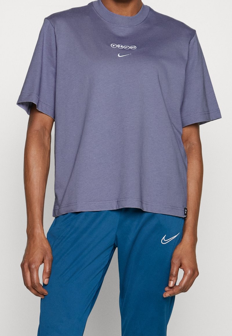 Homme portant un t-shirt Nike lavande ample avec un design circulaire au-dessus du logo et un pantalon de sport Nike bleu.