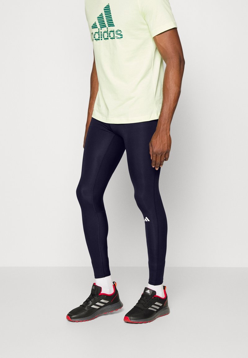 Marina leggings, åtsittande design med vitt Adidas-logotyp; tillsammans med ljusgrön T-shirt med mörkgrön Adidas-text och svarta träningsskor.