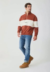 Uomo che indossa un felpa a mezza zip color ruggine e bianco, jeans blu e stivali marroni con lacci, in piedi con una gamba leggermente piegata.