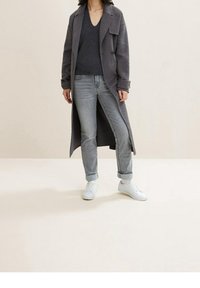 Abrigo largo gris sobre un suéter de cuello en v gris oscuro, combinado con jeans gris claro y zapatillas blancas, presentando un ajuste relajado y puños enrollados.