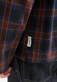 Karierte Hemd, das Nuancen von Braun, Blau und Schwarz zeigt, mit einem Stoffetikett, das "ORIGINALS" beschriftet ist. Die Textur wirkt weich, mit sichtbaren Nähdetails.