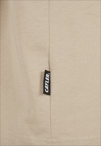 Vaahto beige, jolla on sileä pinta, sisältää mustan etiketin sivussa, jossa lukee "CAYLER". Saumojen yksityiskohdat ovat näkyvissä etiketin lähellä.