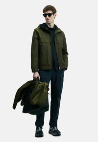 Veste verte imperméable avec deux poches avant, portée avec un pantalon sombre, portant une veste pliée. Des baskets noires complètent la tenue.