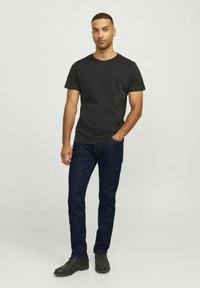 Schwarzes Baumwoll-T-Shirt mit kurzen Ärmeln, kombiniert mit dunkelblauen Jeans. Das Modell steht in schwarzen Schuhen vor einem hellen Hintergrund.