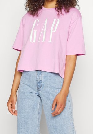Woman wearing a loose pink cropped T-shirt with "GAP" in white letters and light blue high-waisted jeans, standing against a plain background.