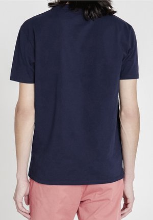 Person mit mittellangen dunklen Haaren, die ein schlichtes dunkelblaues T-Shirt und pinke Hose trägt, von hinten gezeigt.