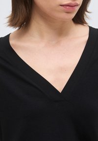 Γυναίκα που φοράει μαύρη μπλούζα με V λαιμόκοψη, εμφανίζοντας τον λαιμό και τις κλειδώσεις της, με κοντά καστανά μαλλιά και φυσικό γυαλιστικό για τα χείλη.