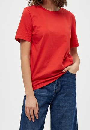 Persoon die een effen rood T-shirt met korte mouwen en een blauwe spijkerbroek draagt, met één hand in de zak en de andere ontspannen langs het lichaam.