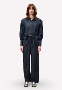 Jumpsuit long à rayures sombres avec un devant à boutons, des manches larges, une ceinture à nouer et une poche poitrine, associé à des chaussures argentées.