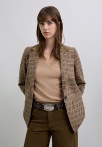 Blazer marrón a cuadros con cuello de ante, combinado con un suéter beige y pantalones de pana marrones. Presenta una hebilla de cinturón decorativa plateada.