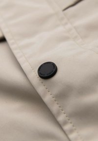 Nahaufnahme von beigem Stoff mit einem schwarzen Knopf, der mit dem Wort "OMBRE" eingraviert ist, das um seinen Rand wiederholt wird.