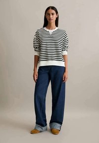 Donna in piedi con una maglietta a maniche lunghe bianca e nera a righe, jeans larghi blu scuro con risvolto e scarpe beige, davanti a uno sfondo neutro.