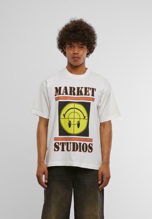 Hvit bomulls t-skjorte med dristig svart og gul grafikk med et smilende ansikt og teksten "MARKET STUDIOS" i oransje og svarte detaljer.