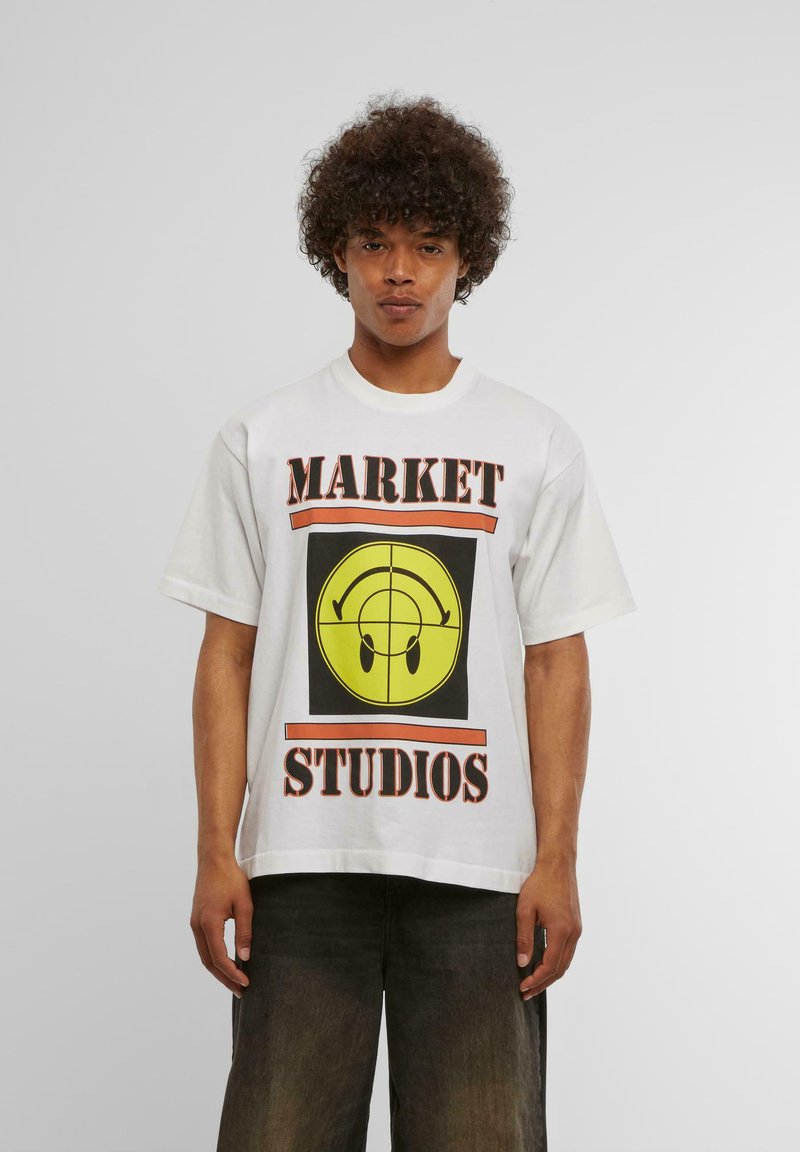 Λευκό βαμβακερό t-shirt με έντονα μαύρα και κίτρινα γραφικά που απεικονίζουν μια χαμογελαστή φατσούλα και το κείμενο "MARKET STUDIOS" με πορτοκαλί και μαύρες λεπτομέρειες.