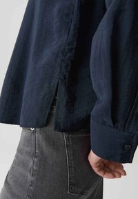 Persona che indossa una camicia a maniche lunghe blu scuro con bottoni ai polsini e pantaloni in denim grigio, mano rilassata lungo il fianco, sfondo in studio.