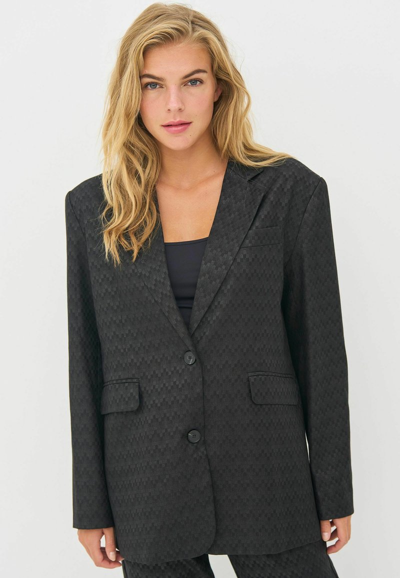 Black patterned blazer with a notched lapel, single button closure, and two front pockets. Made of textured fabric, slightly oversized fit.