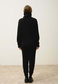 Musta neulepusero, jossa on korkea kaulus, yhdistettynä samansävyisiin mustiin housuihin. Pehmeä rakenne sisältää raidoitetut hihansuut ja helman, mikä tekee siitä sopivan arkikäyttöön.