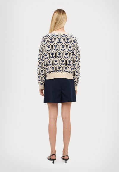 Pull en mâche avec motif fleuri bleu marine, en beige, avec des poignets et un ourlet côtelés. Associé à un short bleu marine, vue de dos visible avec une texture lisse.
