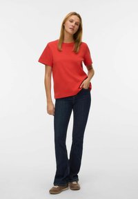 Rød t-shirt med rund hals i bomuldsblanding, korte ærmer, kombineret med mørkeblå, udvidede jeans. Modellen står med den ene hånd i lommen.