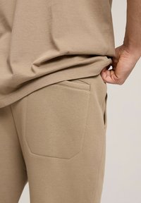 Beżowe bawełniane joggery o dopasowanym kroju, z pojedynczą kieszenią z tyłu i gładką fakturą. Pas jest elastyczny dla większego komfortu.