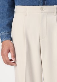 Pantaloni beige su misura con una texture liscia, frontale plissettato e tasche laterali. Una camicia di denim è parzialmente visibile sopra.