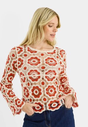 Mujer con una blusa de crochet color crema de manga larga con patrones florales naranjas y jeans azules, con las manos en los bolsillos, mirando hacia un lado.