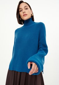 Blauer gerippter Rollkragenpullover mit langen Ärmeln, mit einem gespaltenen Saum und strukturiertem Stoff. Kombiniert mit einem braunen Rock.