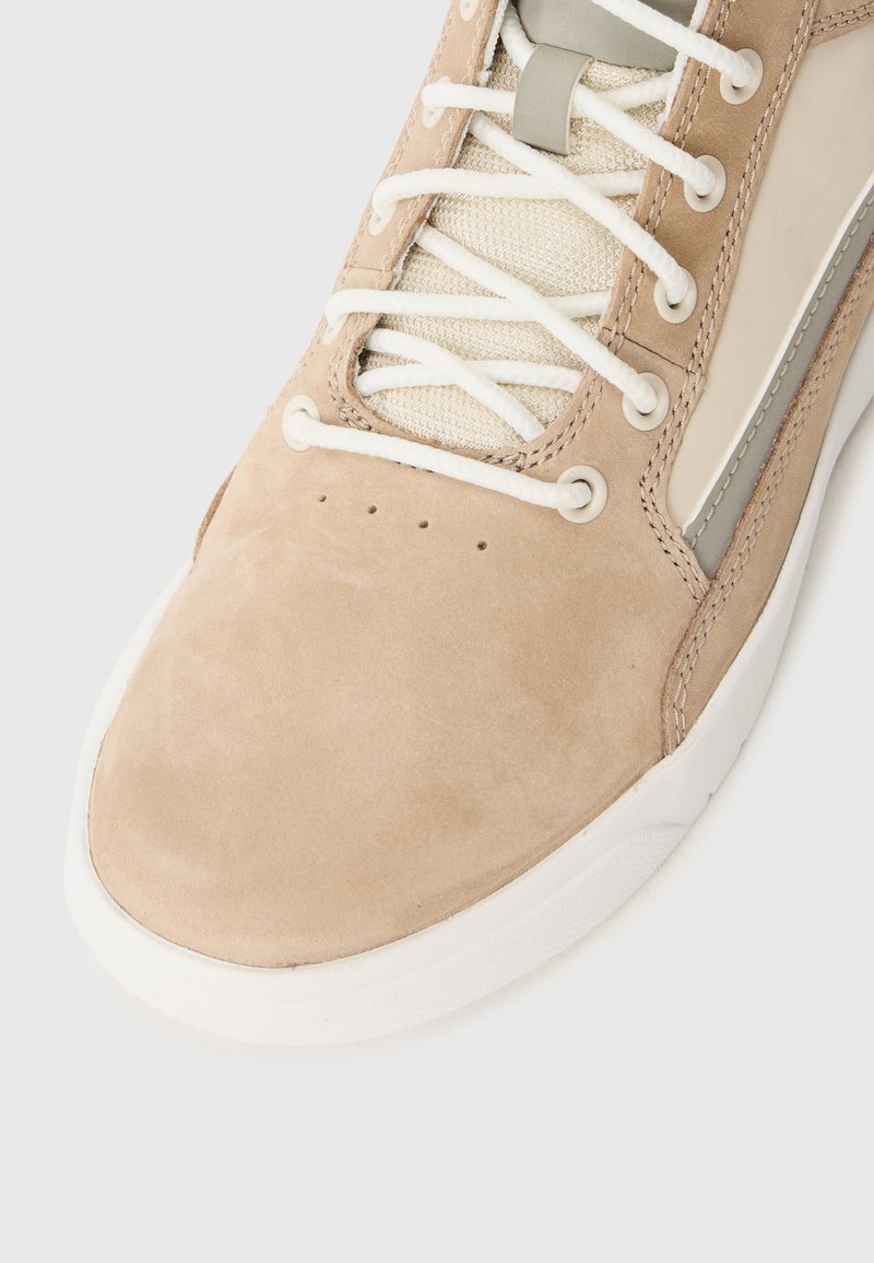 Beige suède sneaker met witte veters en een mesh-tong, voorzien van een lichtgrijze zijaccent en een gestructureerd oppervlak, met een ronde neus.