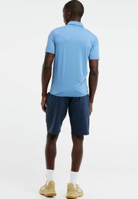 Šviesiai mėlynas, trumpomis rankovėmis polo marškinėliai, lygios medžiagos, derinami su tamsiai mėlynomis šortais su šoninėmis kišenėmis, vilkėti su smėlio spalvos sportinėmis bateliais ir baltais kojiniais.