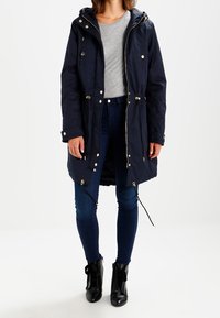 Abrigo largo marine con capucha, cierres de cremallera y botones, cintura ajustable con cordón y detalles en tono dorado. Combinado con jeans oscuros y botines negros.