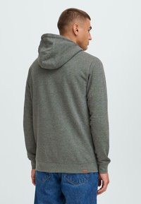 Sweat-shirt à capuche gris avec des manches longues, des poignets et un ourlet côtelés, une texture subtile, et une petite étiquette dans le bas du dos. Porté avec un jean en denim bleu.